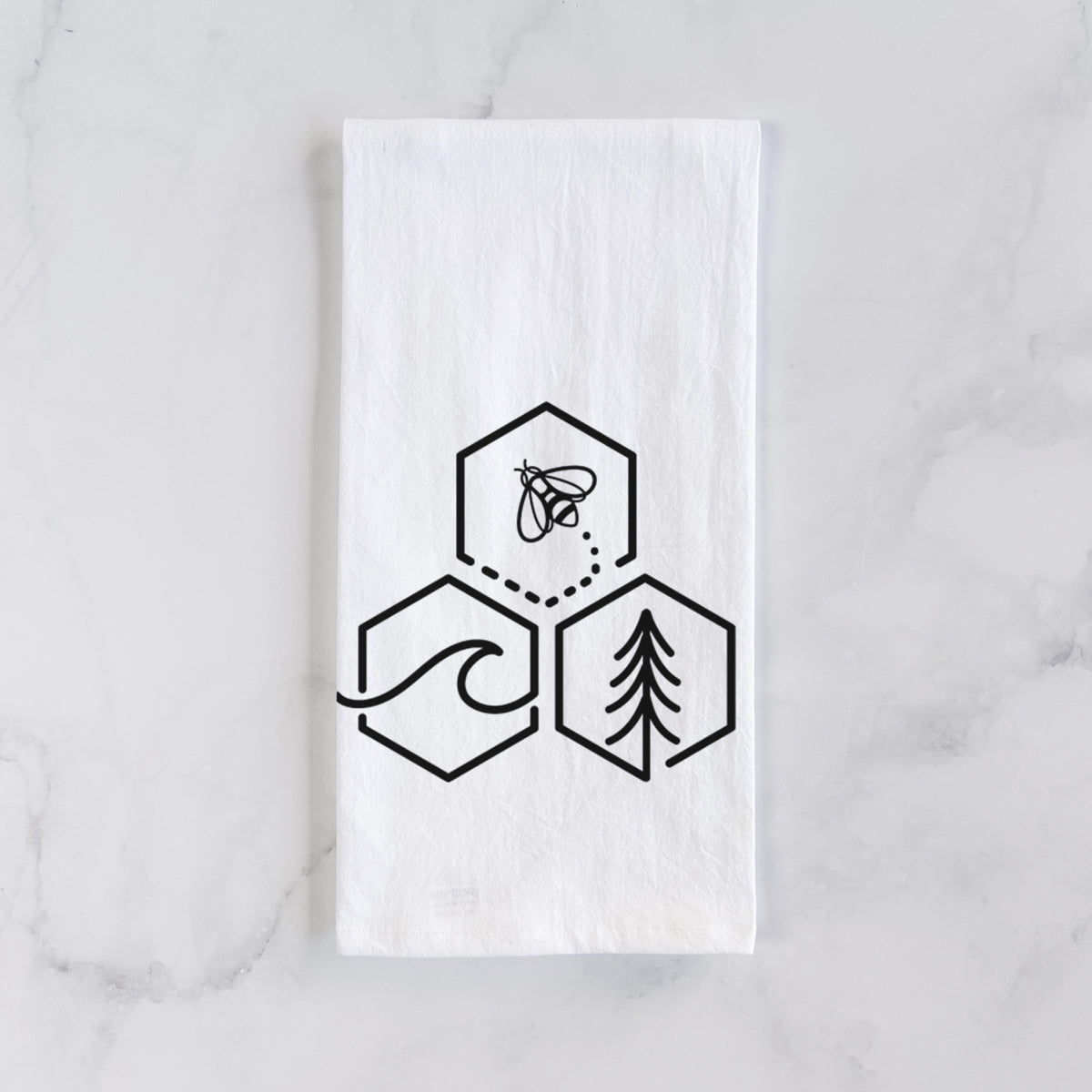 White cotton tea towel laid flat on marble, printed with a black line art design of three hexagons holding a bee, an ocean wave and a pine tree.