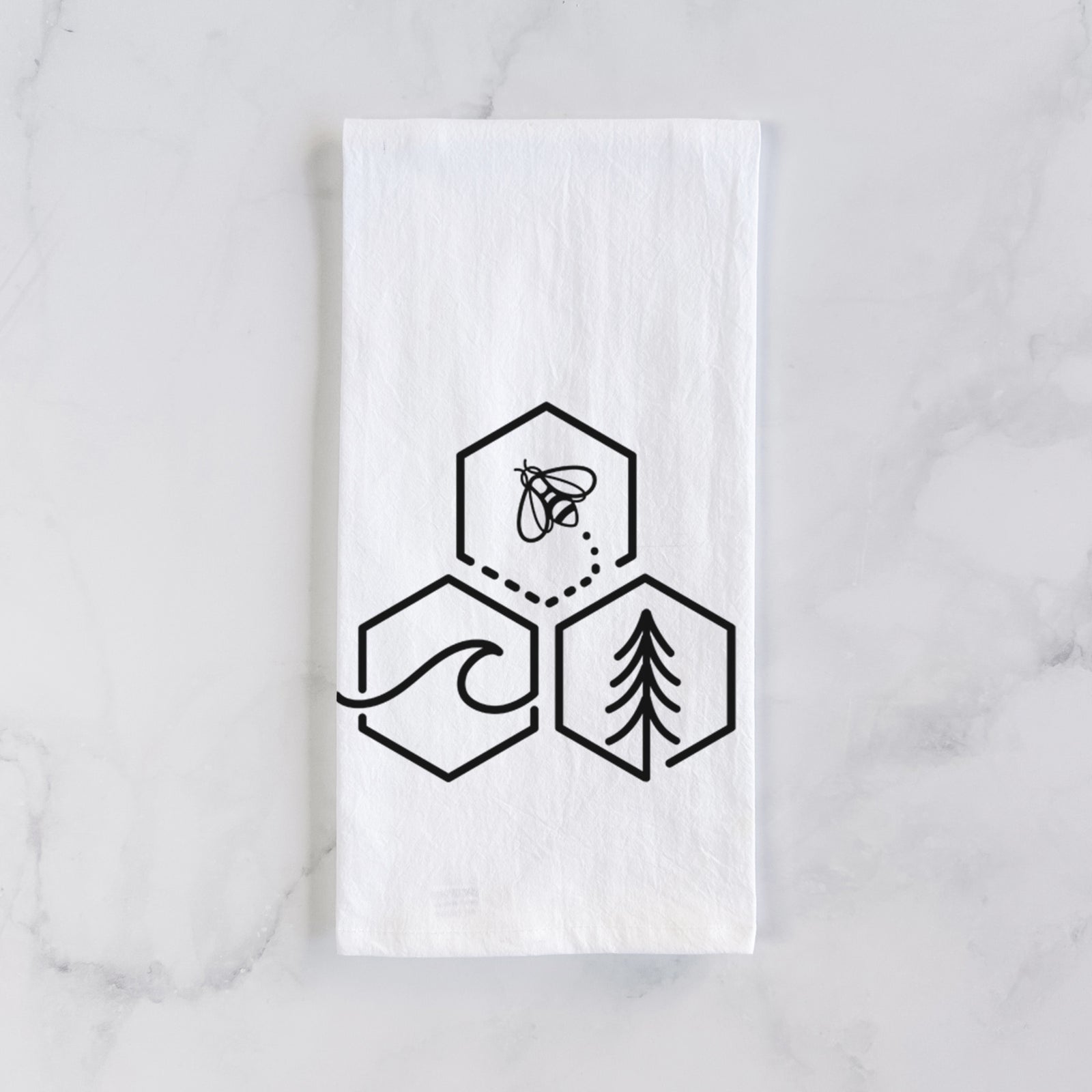 White cotton tea towel printed with a black line art design of three hexagons holding a bee, an ocean wave and a pine tree, on a marble counter beside a round wooden cutting board and greenery.