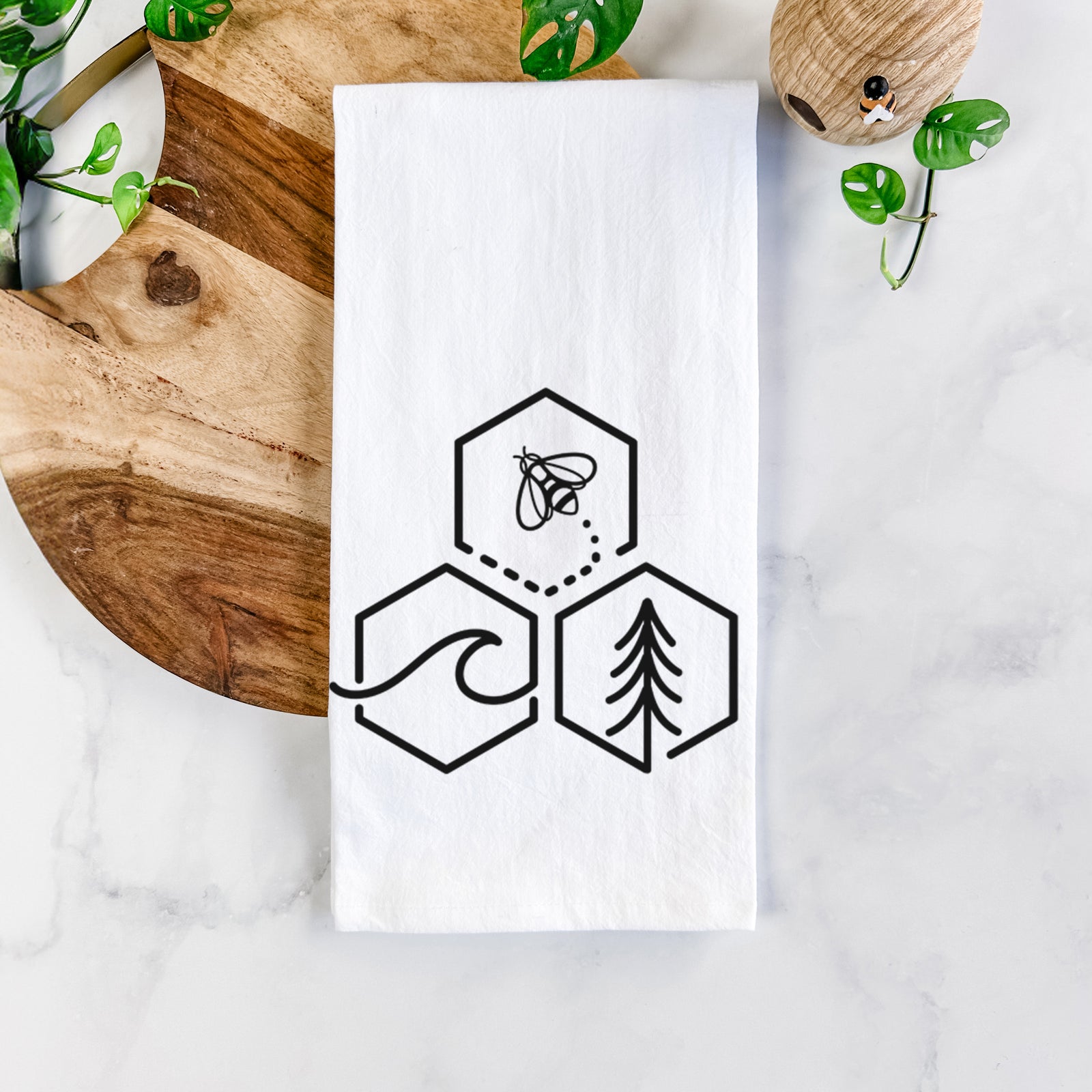 White cotton tea towel printed with a black line art design of three hexagons holding a bee, an ocean wave and a pine tree, on a marble counter beside a round wooden cutting board and greenery.