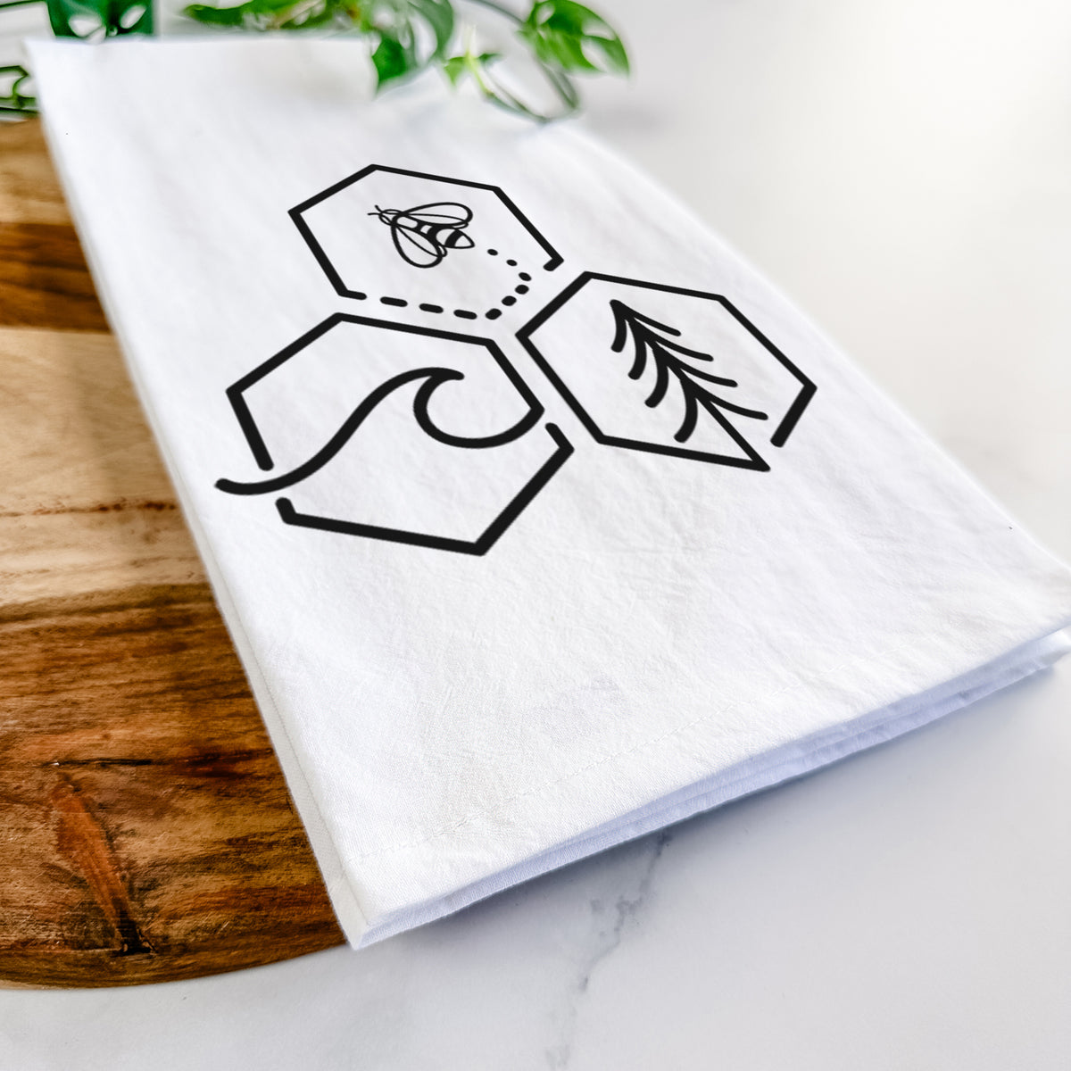 Close-up of a folded white tea towel on a wooden board, showing a black line art design of three hexagons holding a bee, an ocean wave and a pine tree.