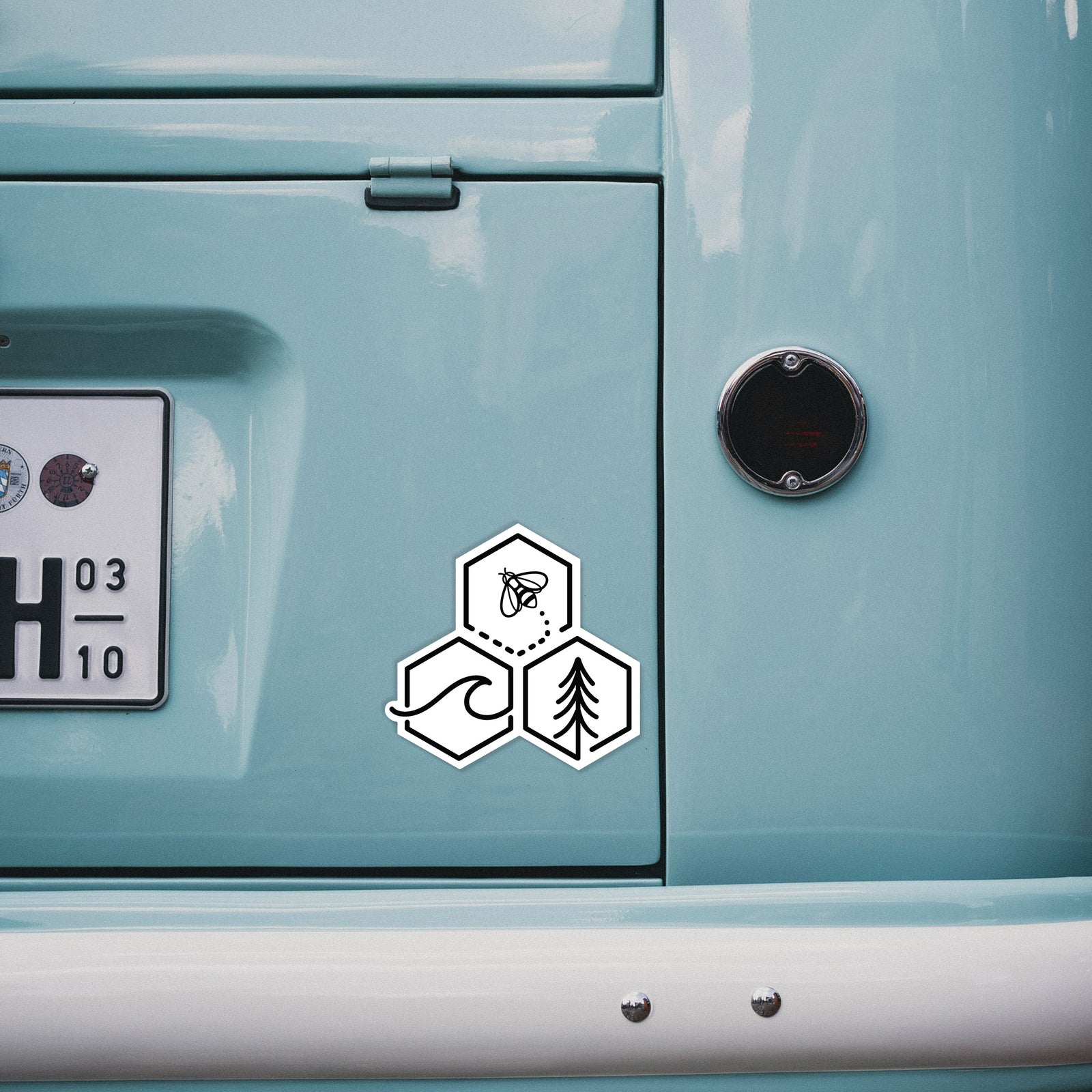Hand holding a 4 inch vinyl decal of a black line art design of three hexagons holding a bee, an ocean wave and a pine tree.