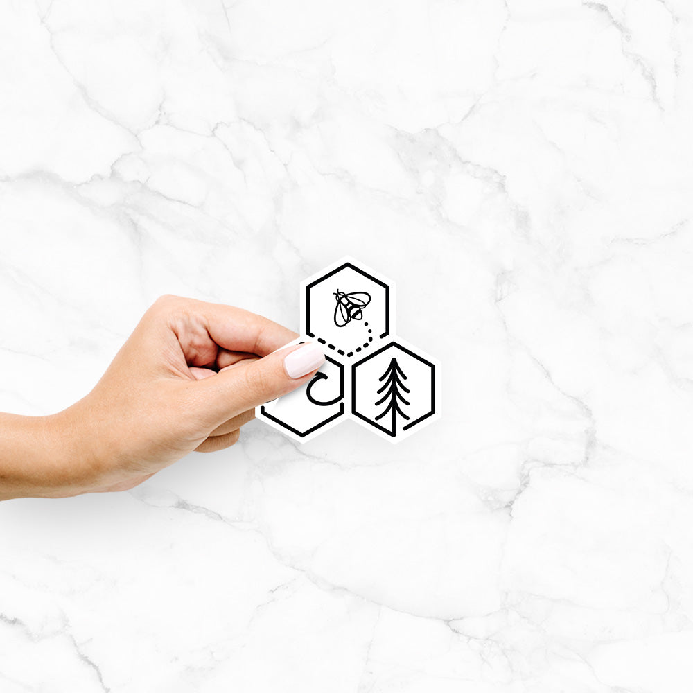 Hand holding a 4 inch vinyl decal of a black line art design of three hexagons holding a bee, an ocean wave and a pine tree.