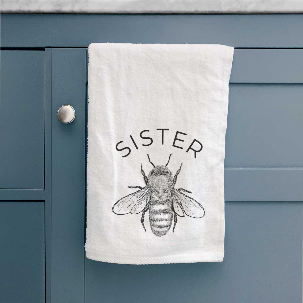 White hand towel printed with a black ink honey bee beneath the word Sister, hanging on a cabinet door.