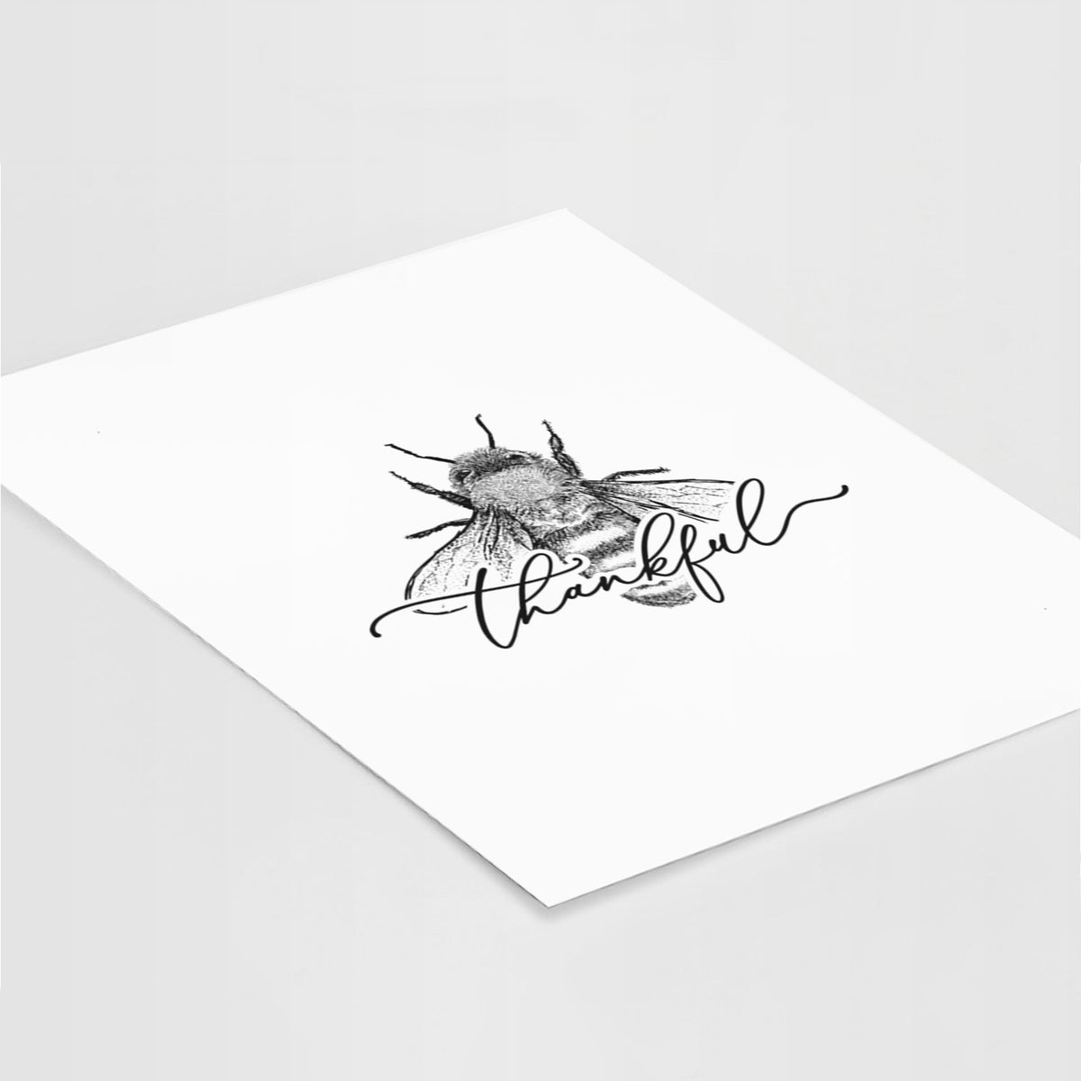 Art print of a black ink bee with the word thankful written in script across it, lying at an angle on a white surface.