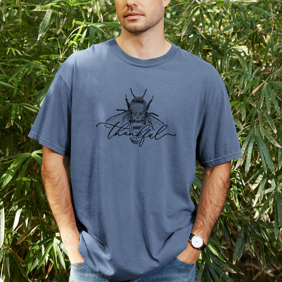 Man wearing a Comfort Colors t-shirt printed with a black ink bee with the word thankful written in script across it, outdoors.