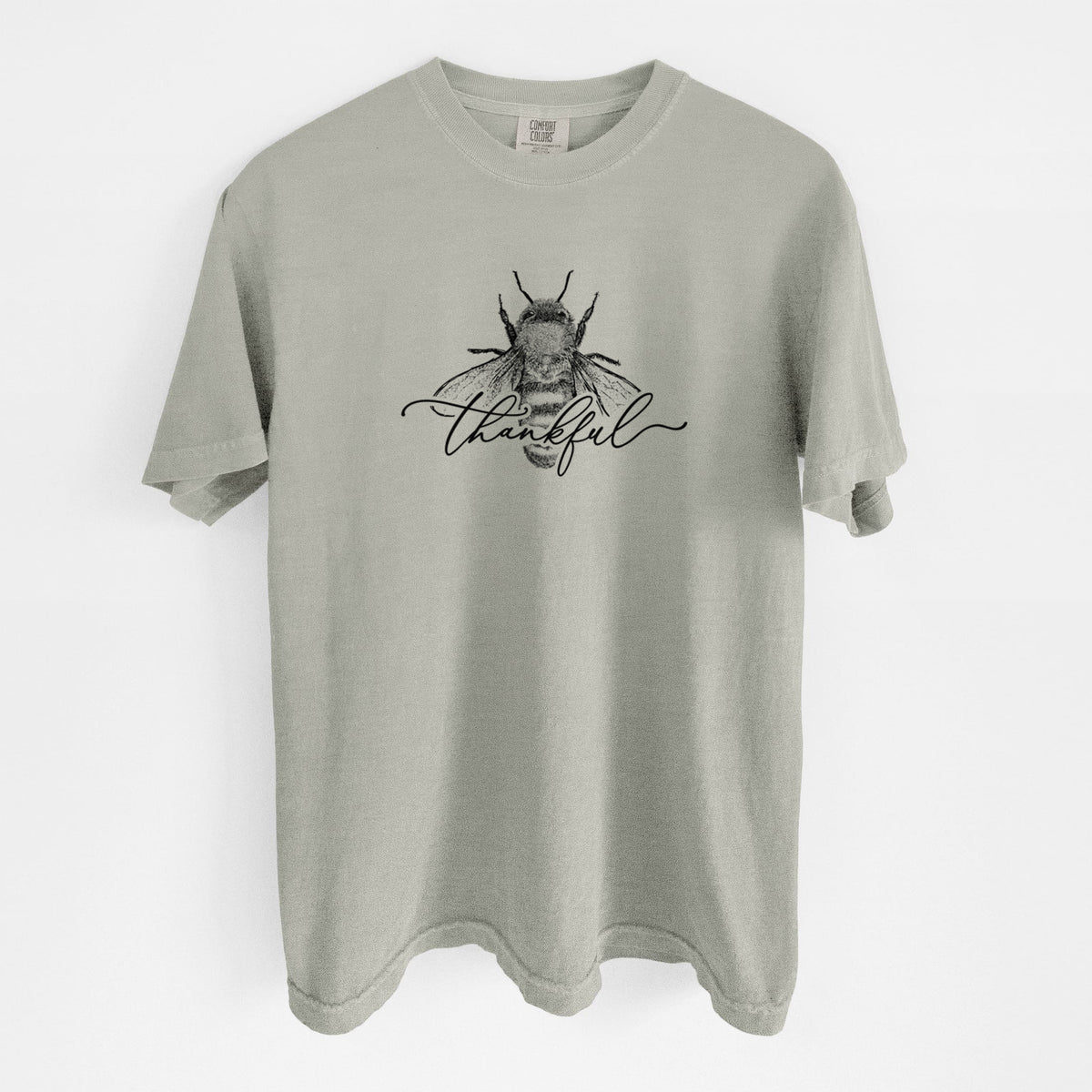 Sandstone beige Comfort Colors t-shirt laid flat, printed with a black ink bee with the word thankful written in script across it.