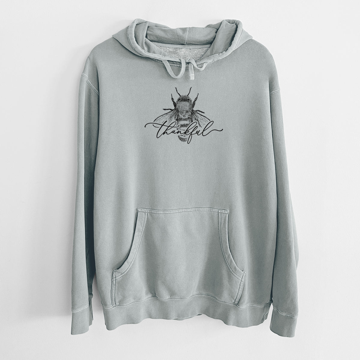 Sage green pigment dyed hoodie laid flat, printed with a black ink bee with the word thankful written in script across it.