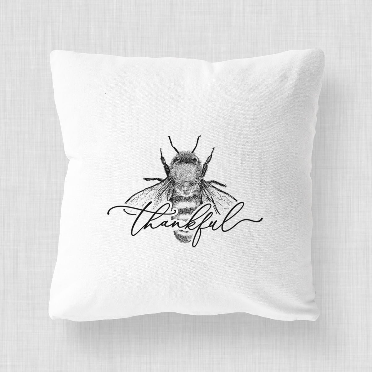 White pillow cover printed with a black ink bee with the word thankful written in script across it.