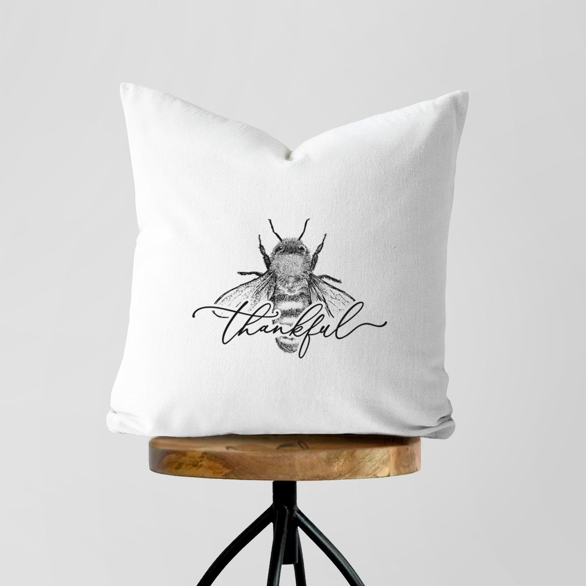White pillow cover printed with a black ink bee with the word thankful written in script across it, on a wooden stool.