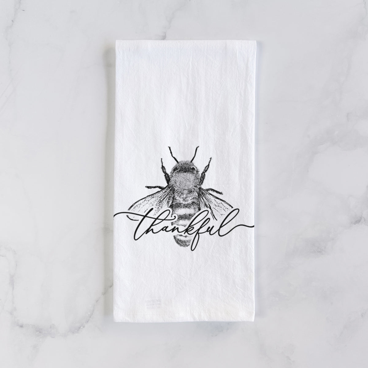 White cotton tea towel laid flat on marble, printed with a black ink bee with the word thankful written in script across it.