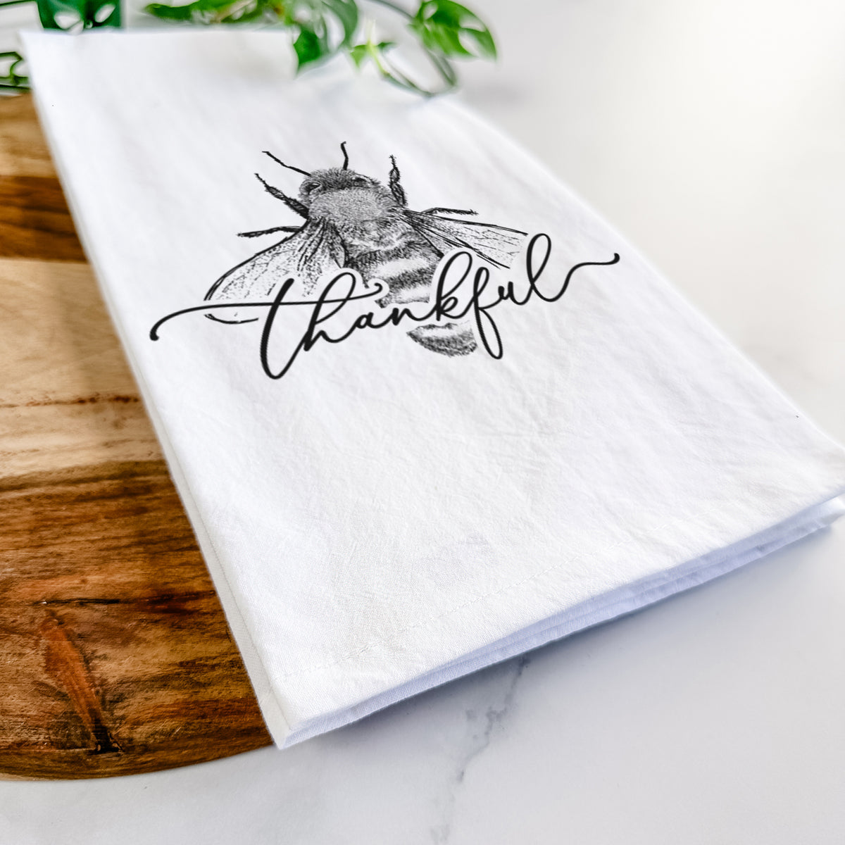 Close-up of a folded white tea towel on a wooden board, showing a black ink bee with the word thankful written in script across it.
