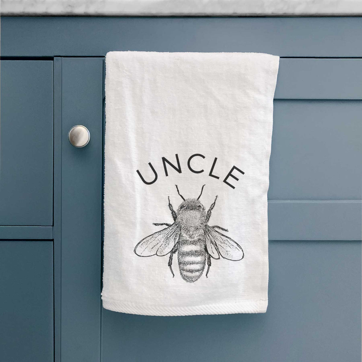 White hand towel printed with a black ink honey bee beneath the word Uncle, hanging on a cabinet door.