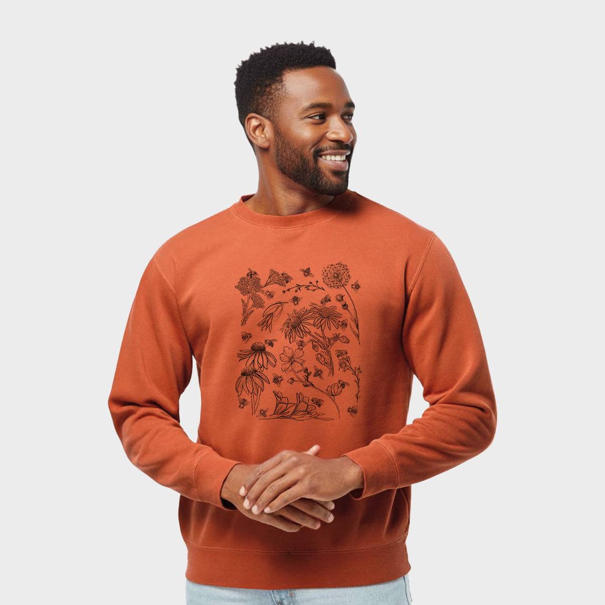 Model wearing an amber orange pigment dyed crewneck sweatshirt printed with black ink bees scattered among wildflowers.