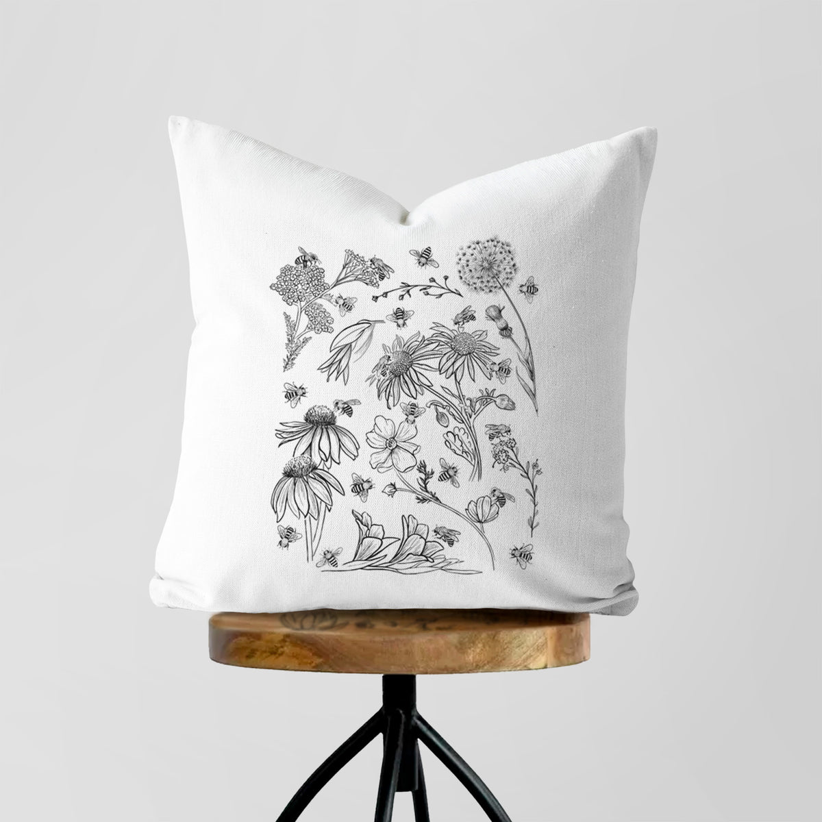 White pillow cover printed with black ink bees scattered among wildflowers, on a wooden stool.