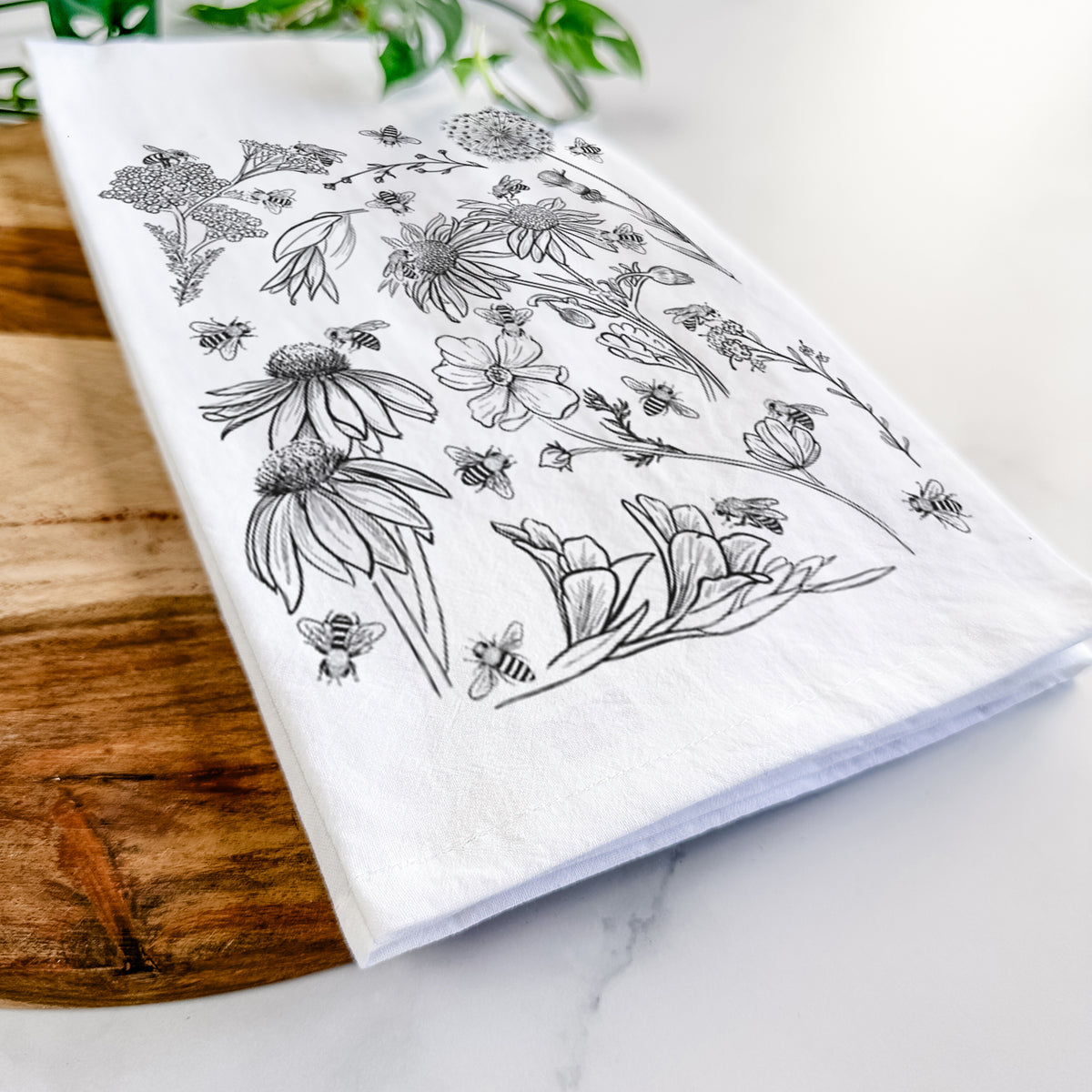 Close-up of a folded white tea towel on a wooden board, showing black ink bees scattered among wildflowers.