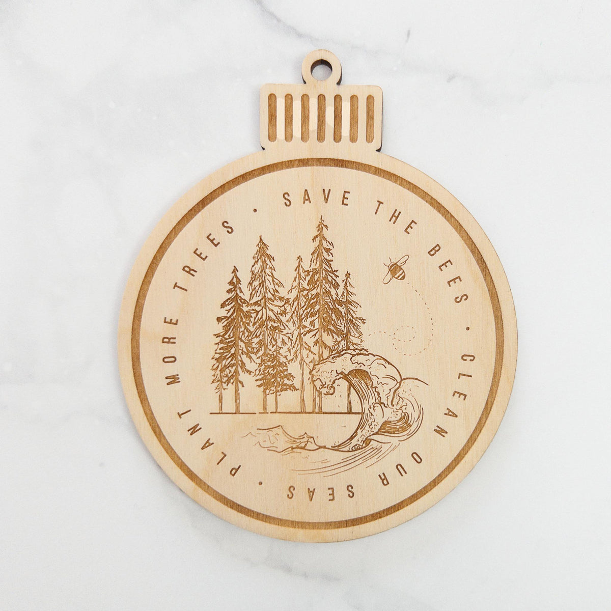 Wooden ornament featuring an engraved round scene of pine trees and an ocean wave ringed by the words Save the Bees, Clean Our Seas, Plant More Trees, on a marble surface.