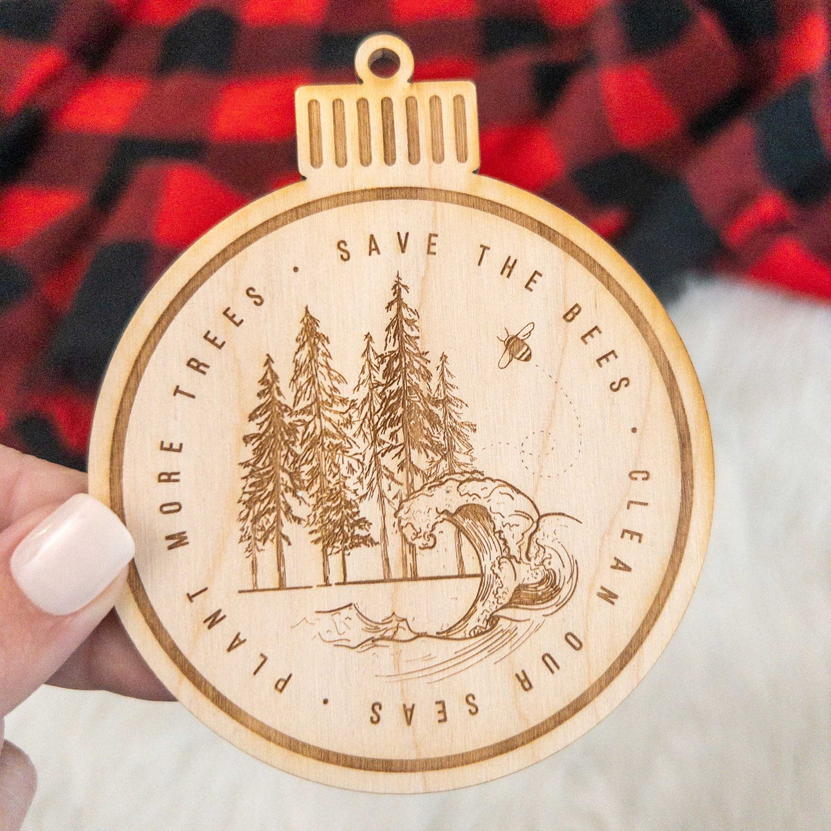 Wooden ornament featuring an engraved round scene of pine trees and an ocean wave ringed by the words Save the Bees, Clean Our Seas, Plant More Trees, on a knit blanket with pine cones.