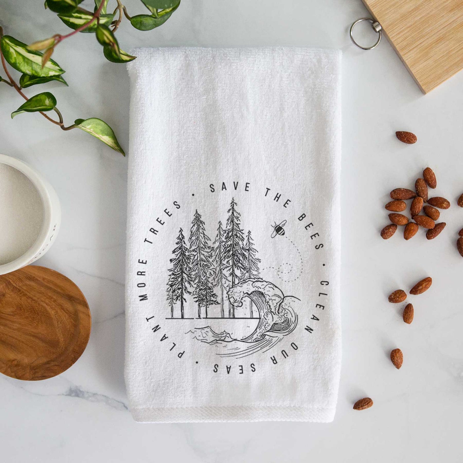 White hand towel printed with a circle of pine trees and an ocean wave ringed by the words Save the Bees, Clean Our Seas, Plant More Trees.