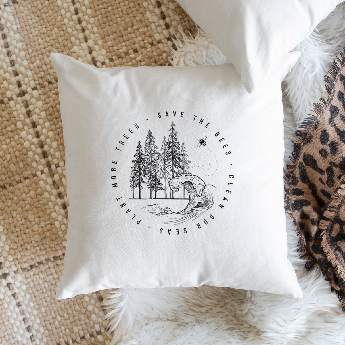 White pillow cover printed with a circle of pine trees and an ocean wave ringed by the words Save the Bees, Clean Our Seas, Plant More Trees, on a woven chair.