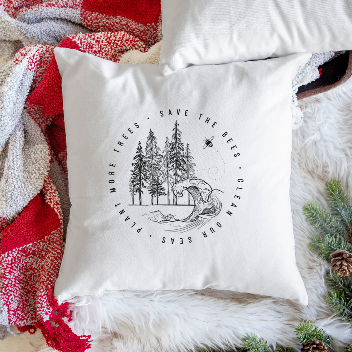 White pillow cover printed with a circle of pine trees and an ocean wave ringed by the words Save the Bees, Clean Our Seas, Plant More Trees, on a woven chair.