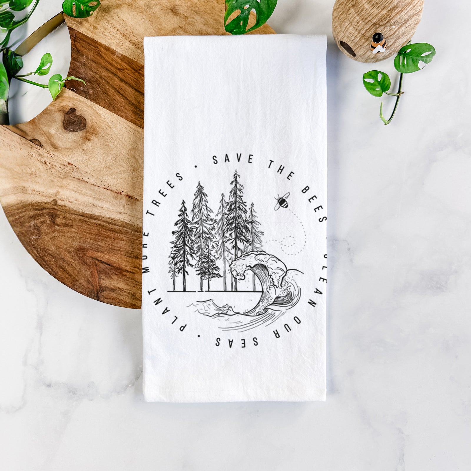 White cotton tea towel printed with a circle of pine trees and an ocean wave ringed by the words Save the Bees, Clean Our Seas, Plant More Trees, on a marble counter beside a round wooden cutting board and greenery.