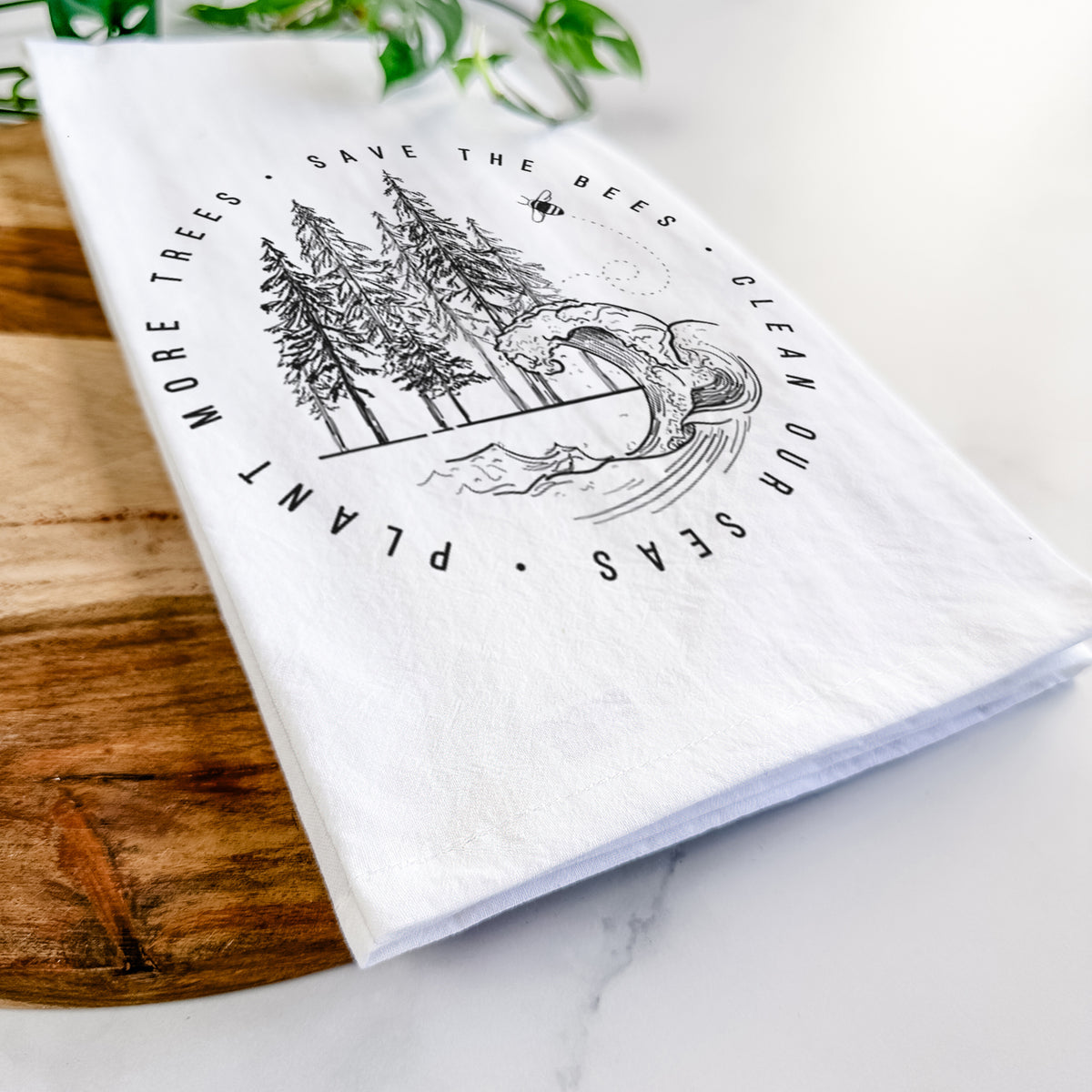Close-up of a folded white tea towel on a wooden board, showing a circle of pine trees and an ocean wave ringed by the words Save the Bees, Clean Our Seas, Plant More Trees.
