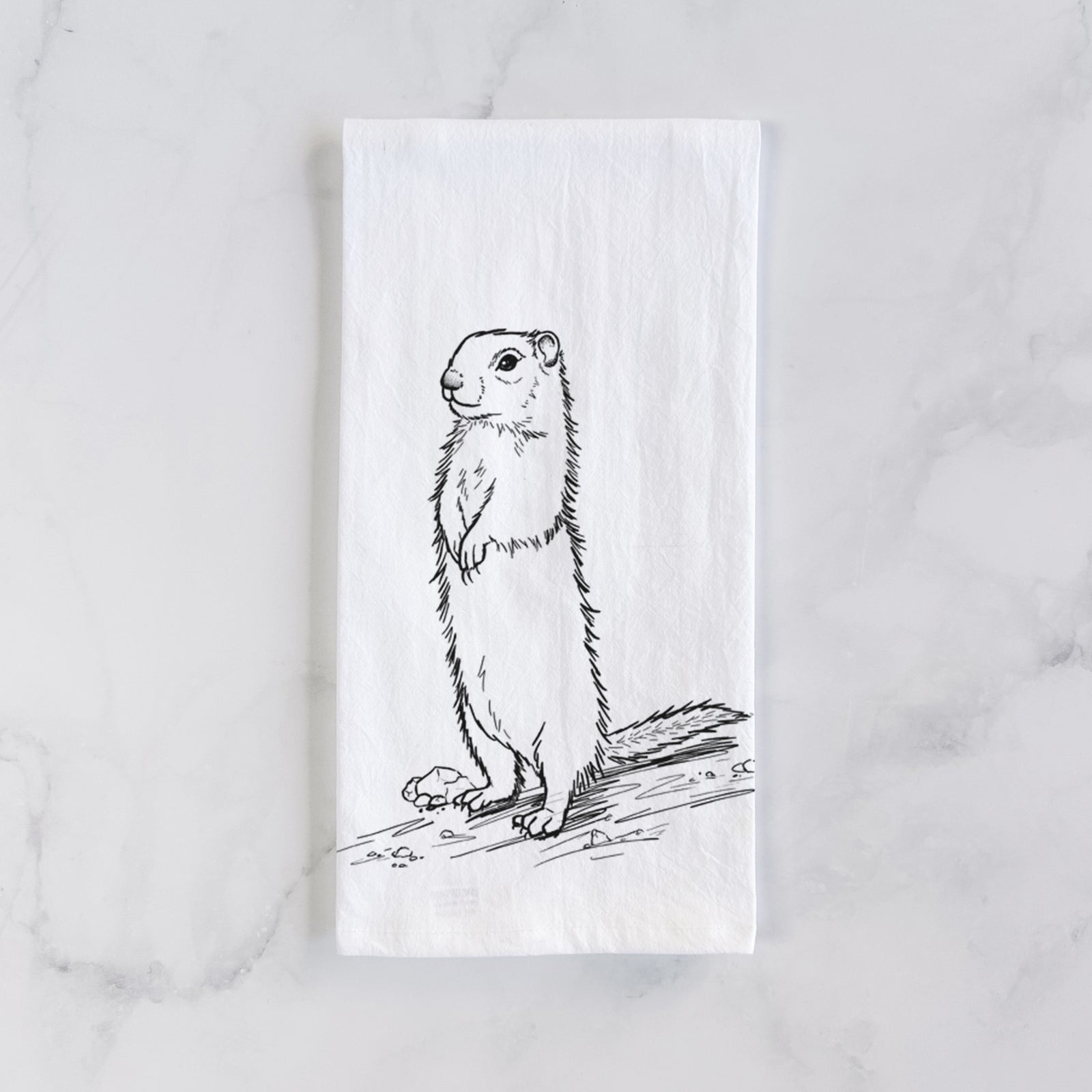 White cotton tea towel printed with a detailed black ink illustration of a Belding's ground squirrel standing upright, on a marble counter beside a round wooden cutting board and greenery.