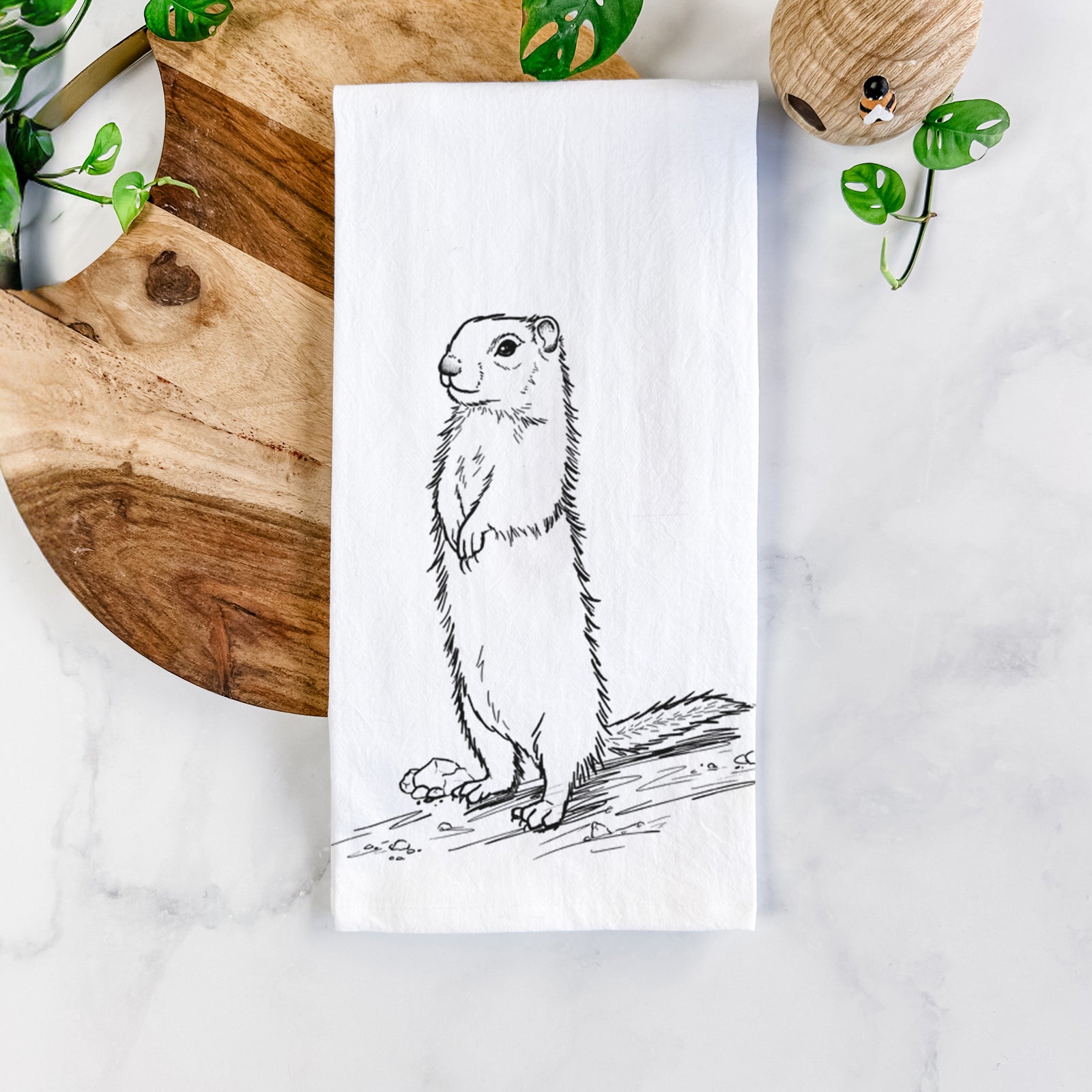 White cotton tea towel printed with a detailed black ink illustration of a Belding's ground squirrel standing upright, on a marble counter beside a round wooden cutting board and greenery.