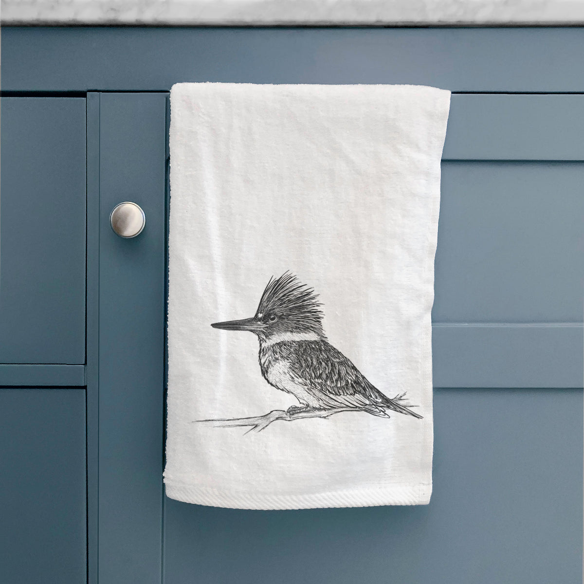 White hand towel printed with a detailed black ink sketch of a belted kingfisher perched on a branch, hanging on a cabinet door.