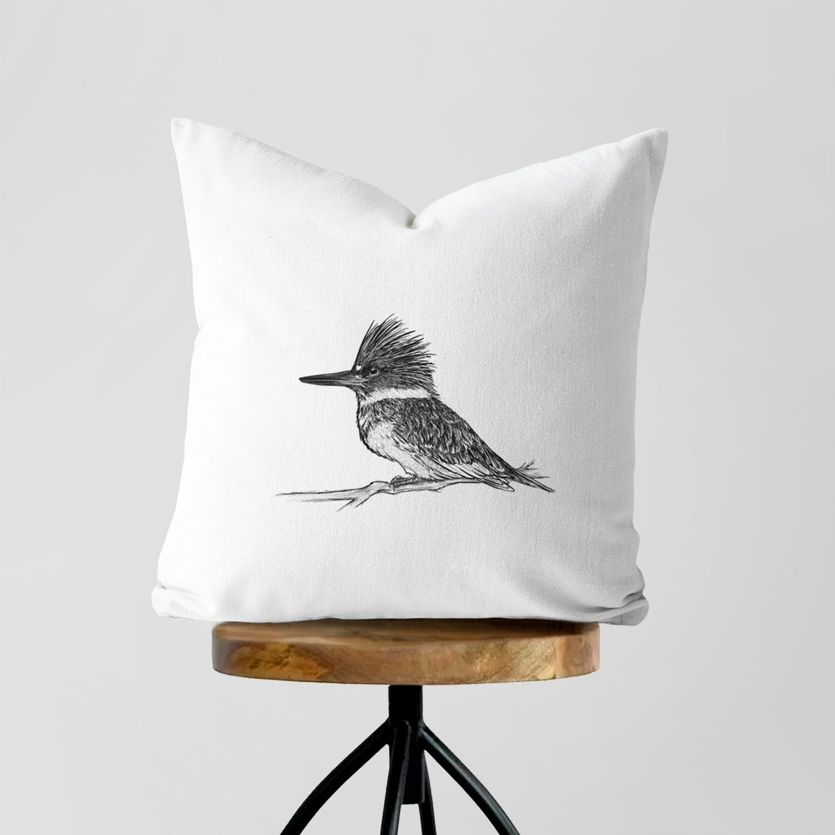 White pillow cover printed with a detailed black ink sketch of a belted kingfisher perched on a branch, on a wooden stool.