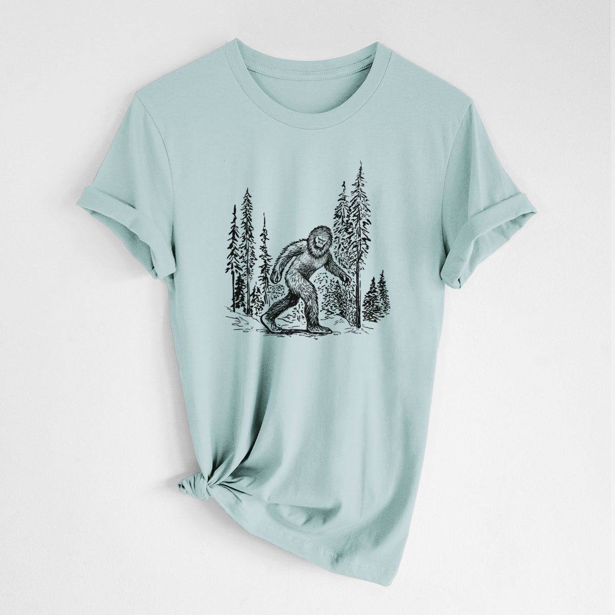 Dusty blue lightweight cotton t-shirt laid flat with the sleeves rolled, printed with a black ink illustration of Bigfoot walking through a pine forest.