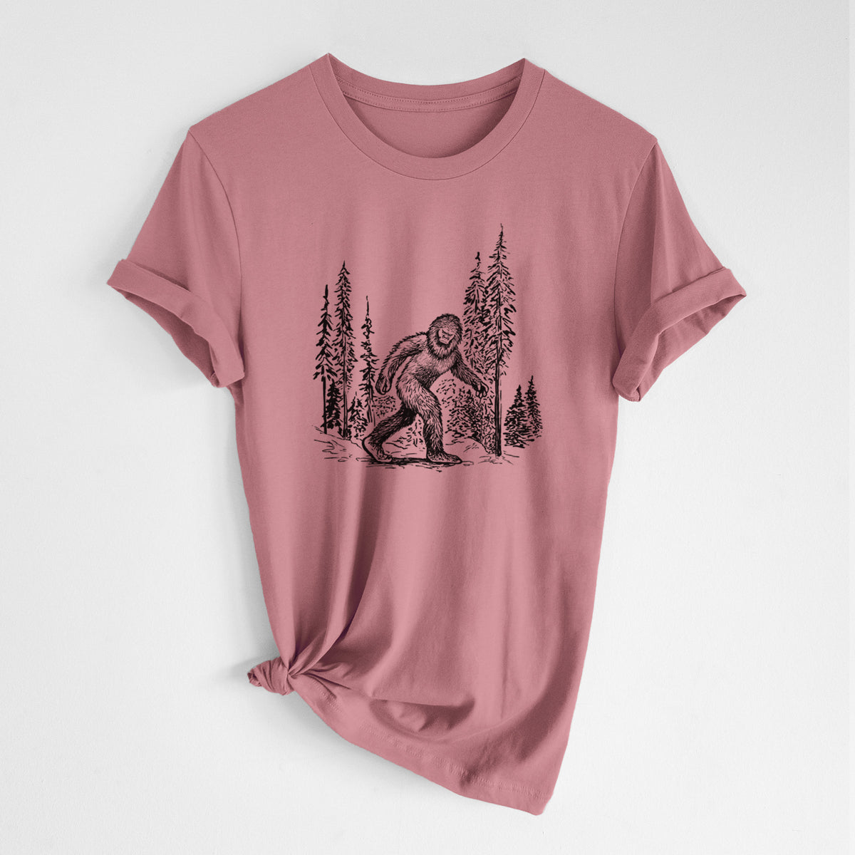 Mauve pink lightweight cotton t-shirt laid flat with the sleeves rolled, printed with a black ink illustration of Bigfoot walking through a pine forest.