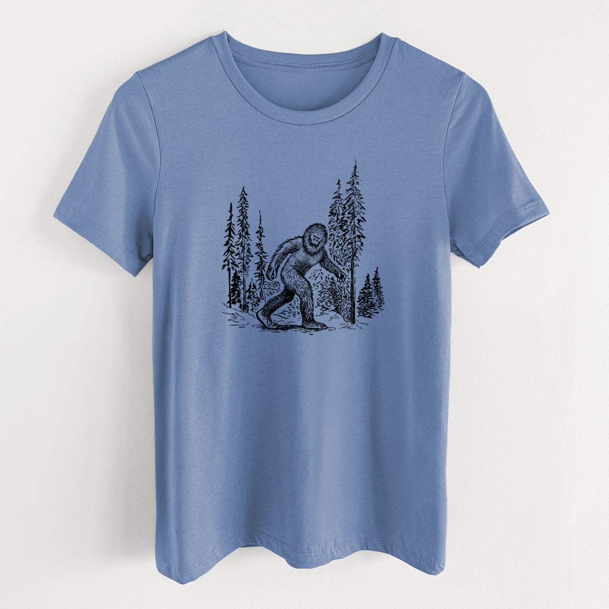 Blue bell women's relaxed fit cotton t-shirt laid flat, printed with a black ink illustration of Bigfoot walking through a pine forest.