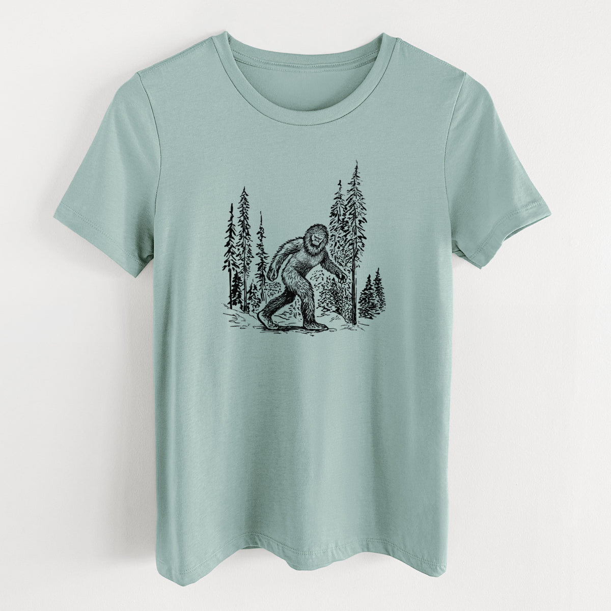 Dusty blue women's relaxed fit cotton t-shirt laid flat, printed with a black ink illustration of Bigfoot walking through a pine forest.