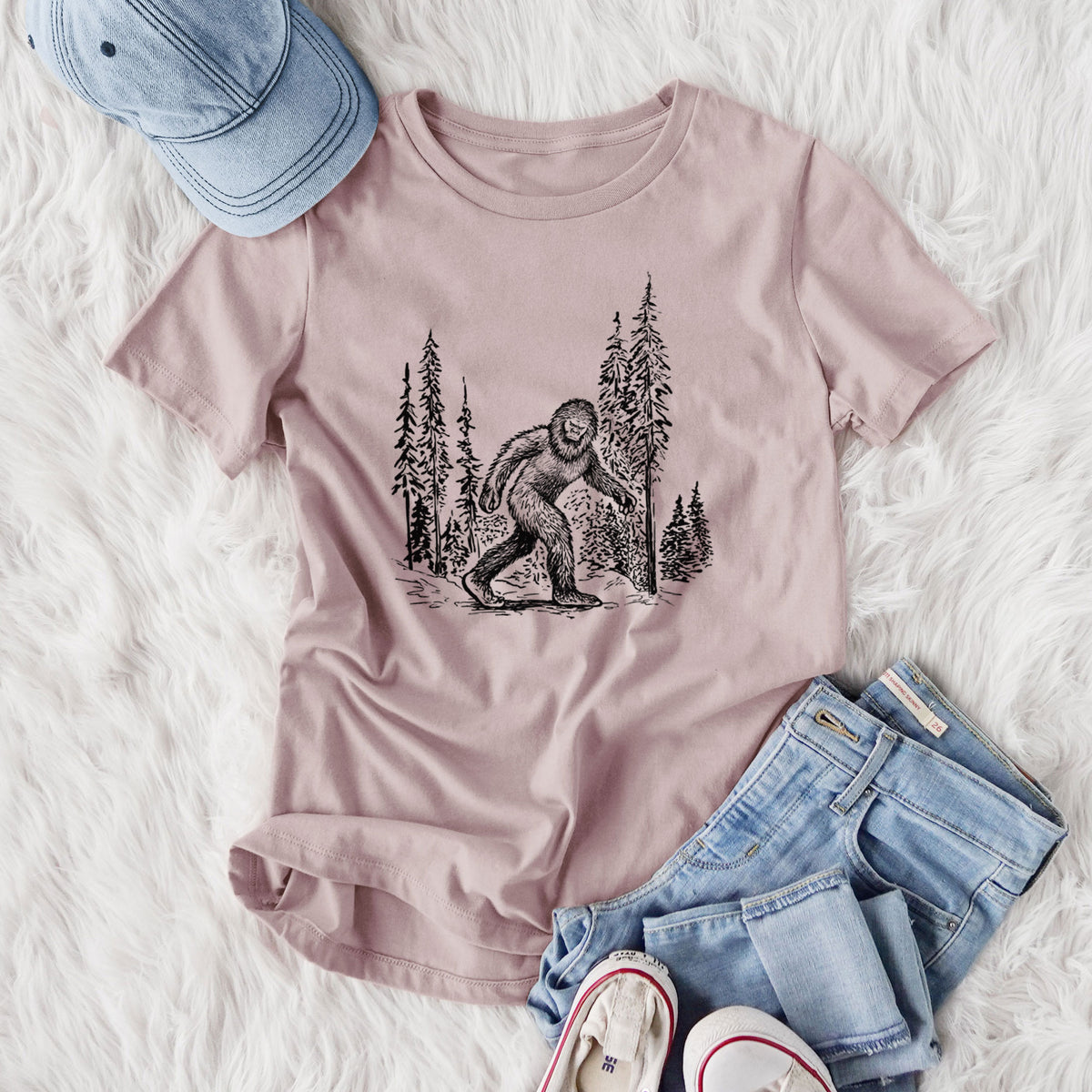 Women's relaxed fit cotton t-shirt printed with a black ink illustration of Bigfoot walking through a pine forest, styled flat on a white faux fur rug with jeans.