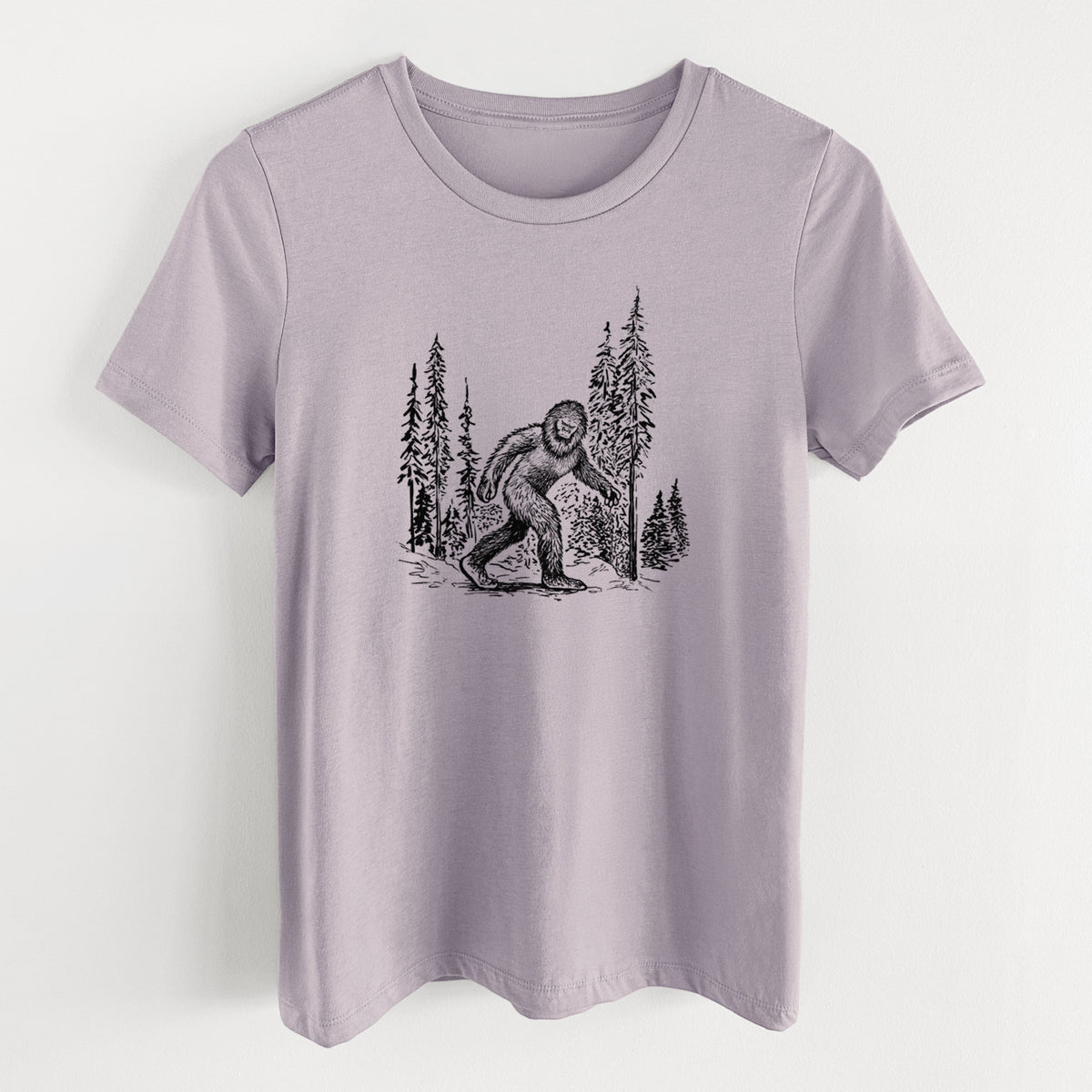 Light violet women's relaxed fit cotton t-shirt laid flat, printed with a black ink illustration of Bigfoot walking through a pine forest.