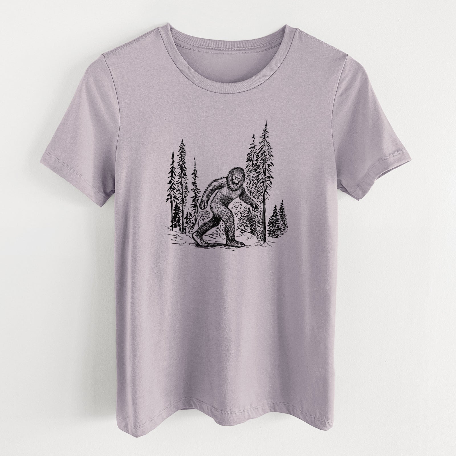 Light violet women's relaxed fit cotton t-shirt laid flat, printed with a black ink illustration of Bigfoot walking through a pine forest.