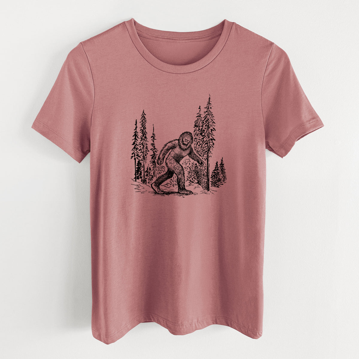 Mauve pink women's relaxed fit cotton t-shirt laid flat, printed with a black ink illustration of Bigfoot walking through a pine forest.