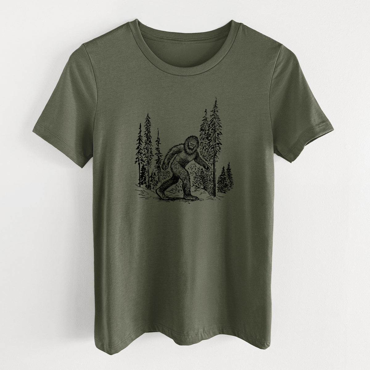 Military green women's relaxed fit cotton t-shirt laid flat, printed with a black ink illustration of Bigfoot walking through a pine forest.