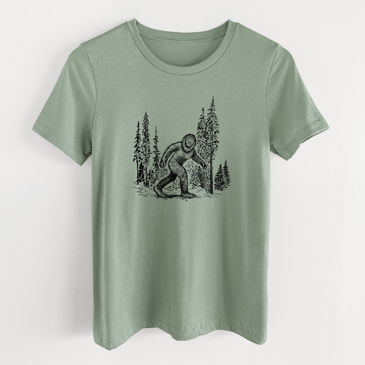 Sage green women's relaxed fit cotton t-shirt laid flat, printed with a black ink illustration of Bigfoot walking through a pine forest.