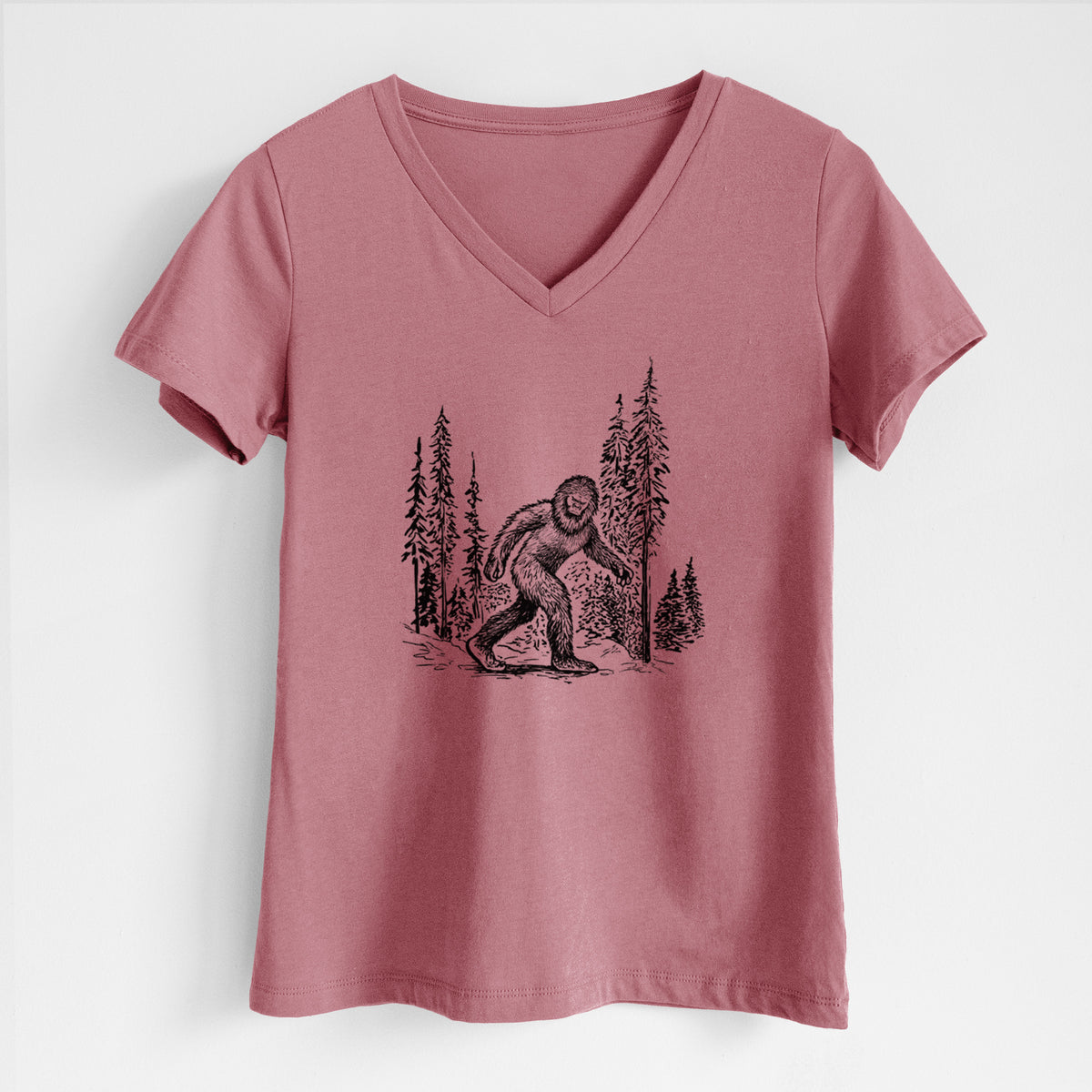 Mauve pink women's relaxed fit cotton V-neck t-shirt laid flat, printed with a black ink illustration of Bigfoot walking through a pine forest.
