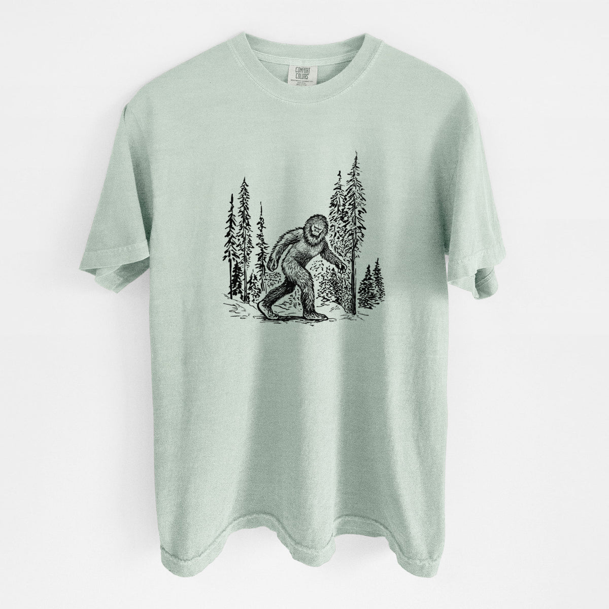 Bay green Comfort Colors t-shirt laid flat, printed with a black ink illustration of Bigfoot walking through a pine forest.