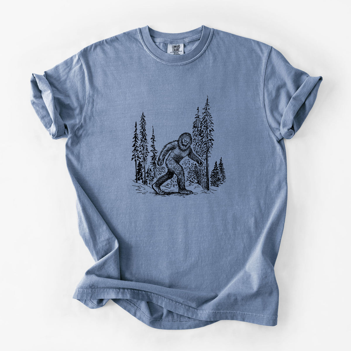 Blue jean Comfort Colors t-shirt laid flat, printed with a black ink illustration of Bigfoot walking through a pine forest.