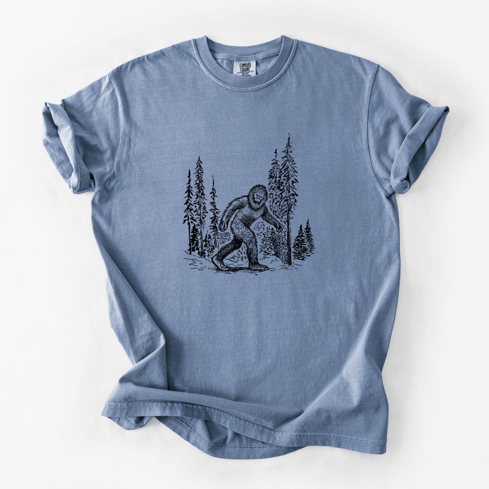 Blue jean Comfort Colors t-shirt laid flat, printed with a black ink illustration of Bigfoot walking through a pine forest.