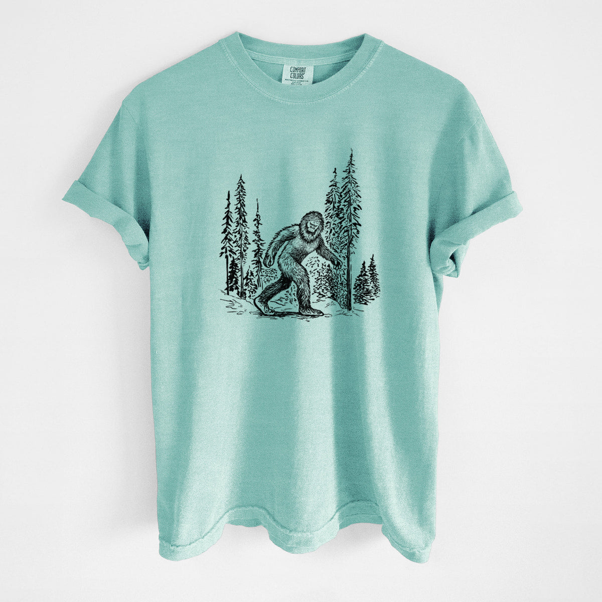Chalky mint Comfort Colors t-shirt laid flat, printed with a black ink illustration of Bigfoot walking through a pine forest.