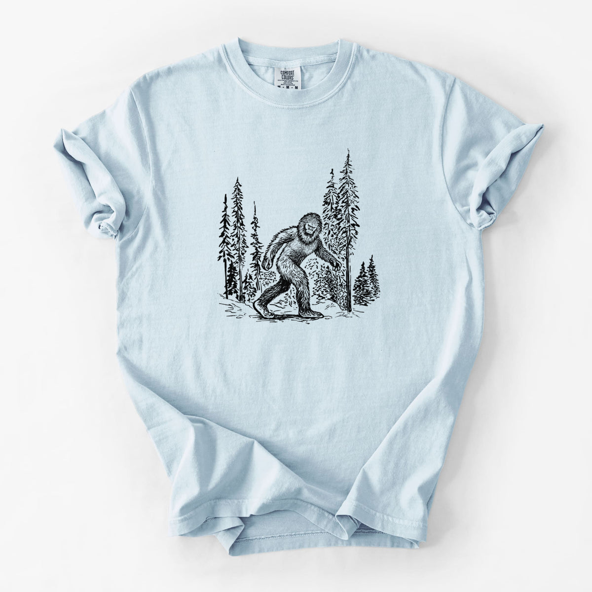 Chambray blue Comfort Colors t-shirt laid flat, printed with a black ink illustration of Bigfoot walking through a pine forest.
