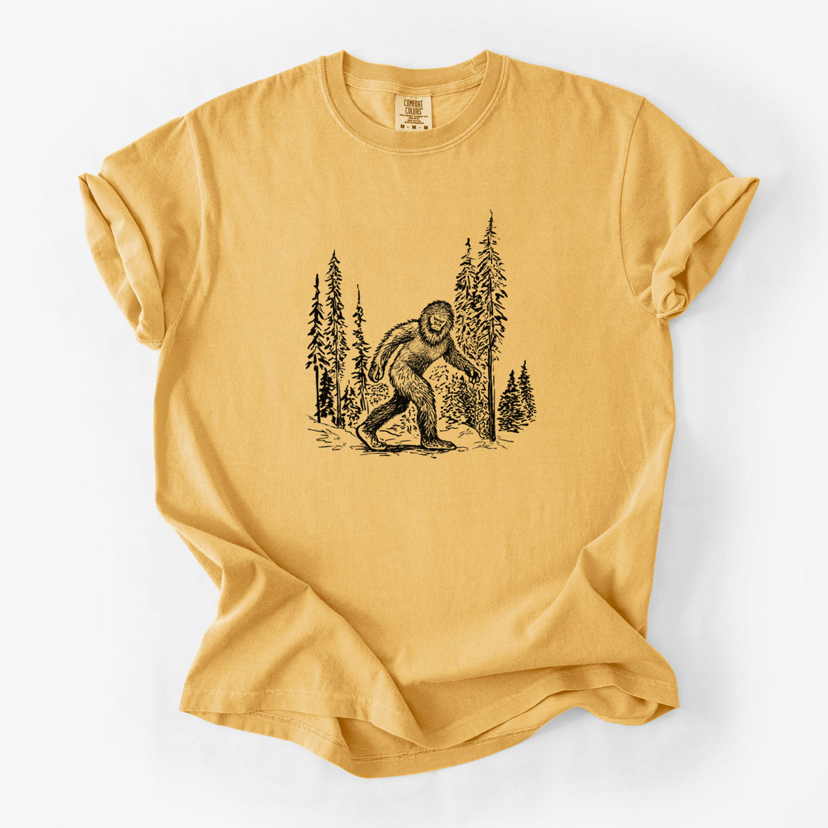 Golden wheat yellow Comfort Colors t-shirt laid flat, printed with a black ink illustration of Bigfoot walking through a pine forest.