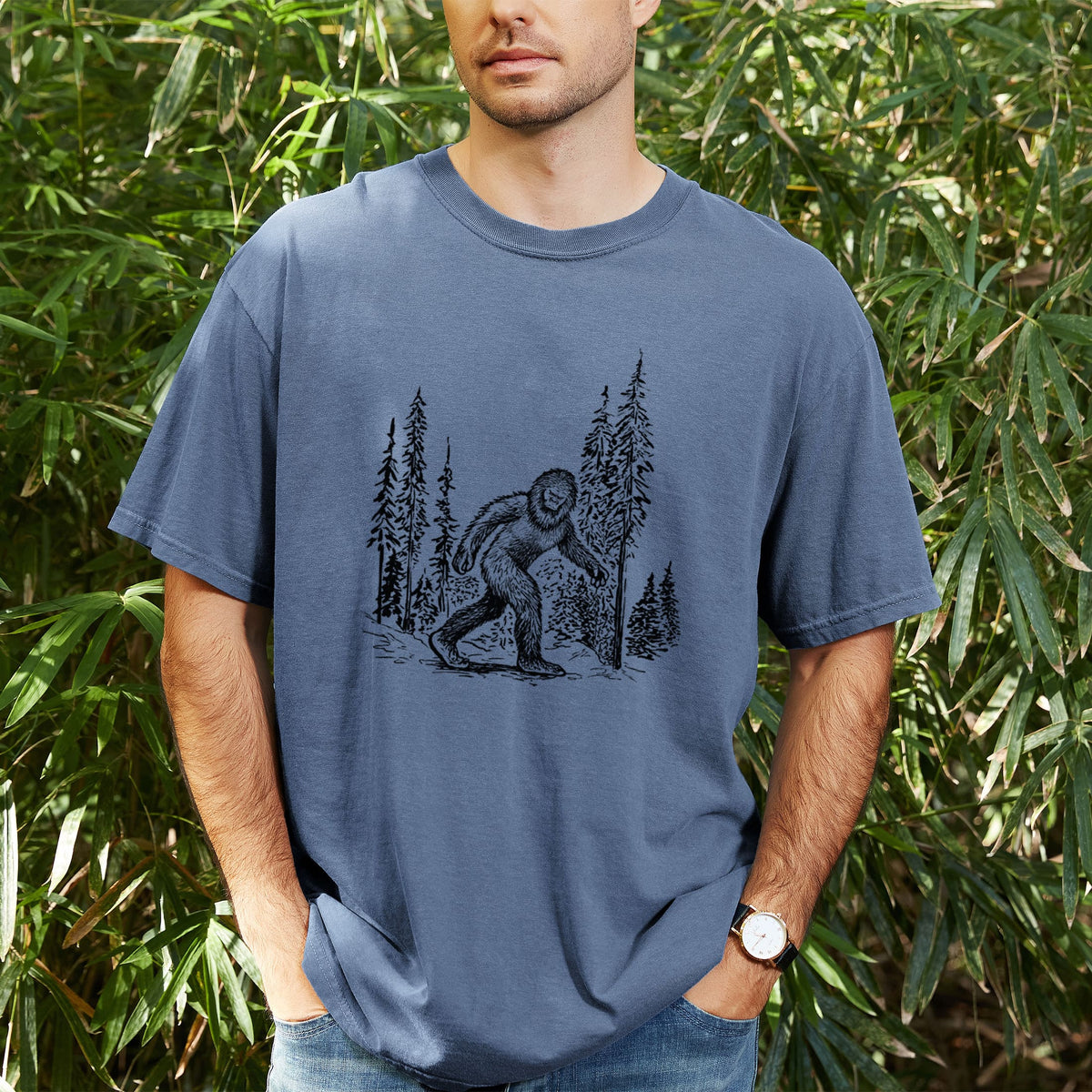 Man wearing a Comfort Colors t-shirt printed with a black ink illustration of Bigfoot walking through a pine forest, outdoors.
