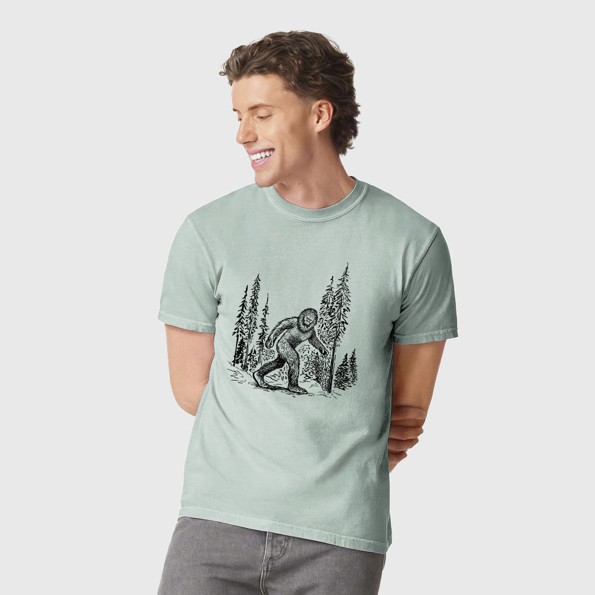Man wearing a Comfort Colors t-shirt printed with a black ink illustration of Bigfoot walking through a pine forest.