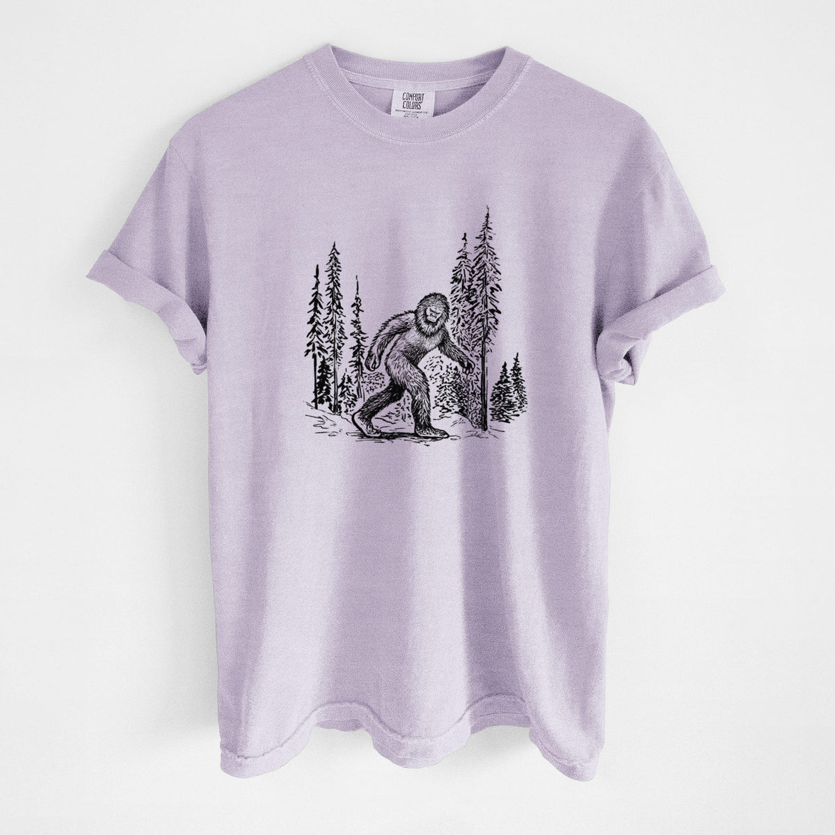 Orchid purple Comfort Colors t-shirt laid flat, printed with a black ink illustration of Bigfoot walking through a pine forest.